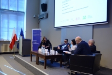 Paneliści siedzą na podium auli na kanapach przed stolikiem.