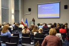 Przy mównicy stoi kobieta i przemawia przed zgromadzonymi na auli uczestnikami