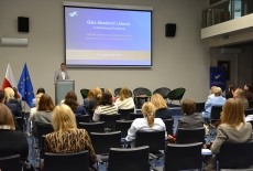 Przy mównicy stoi Zasytępca Dyrektora i przemawia przed zgromadzonymi na auli uczestnikami